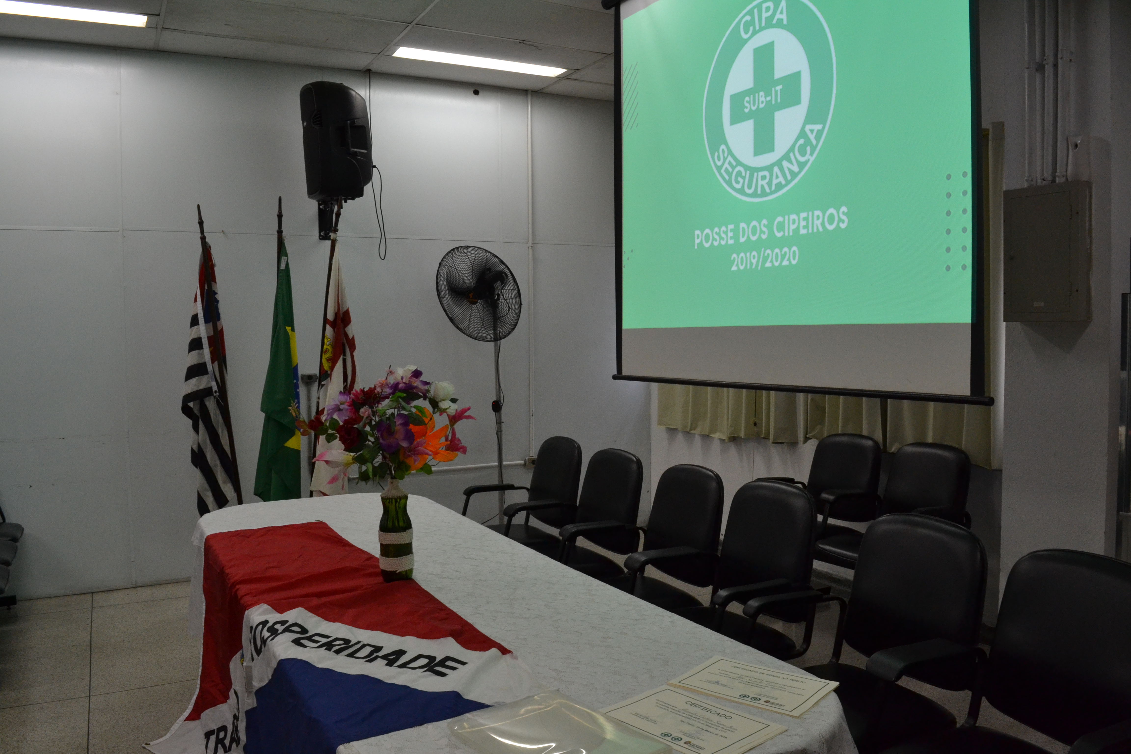 Foto feita em ambiente interno. Nela aparece três bandeiras: do Estado de São Paulo, do Brasil e do Município de São Paulo. A bandeira do Itaim Paulista está sobre uma mesa.