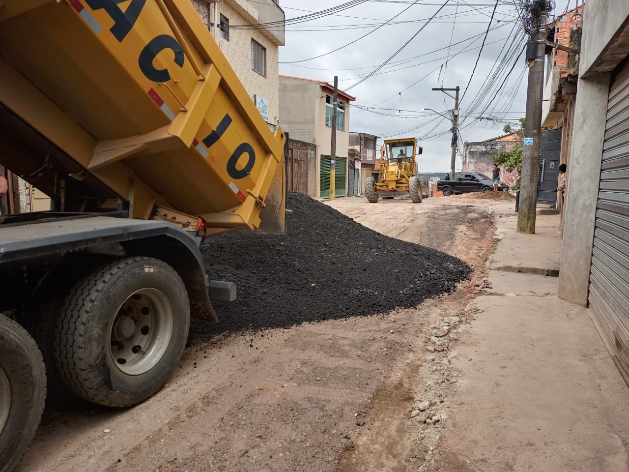Caçamba amarela de um caminhão basculante erguida, despejando fresa de asfalto em uma rua de terra. 