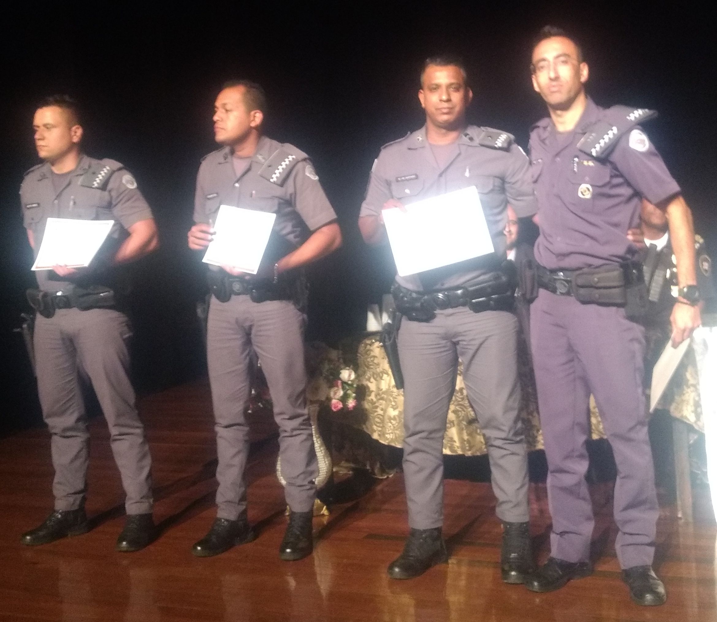 Quatro policiais recebem homenagem pelos trabalhos prestados à população. Estão fardados, em pé, com o certificado à mostra, na altura do peito. É um papel no tamanho de um diploma escolar. 