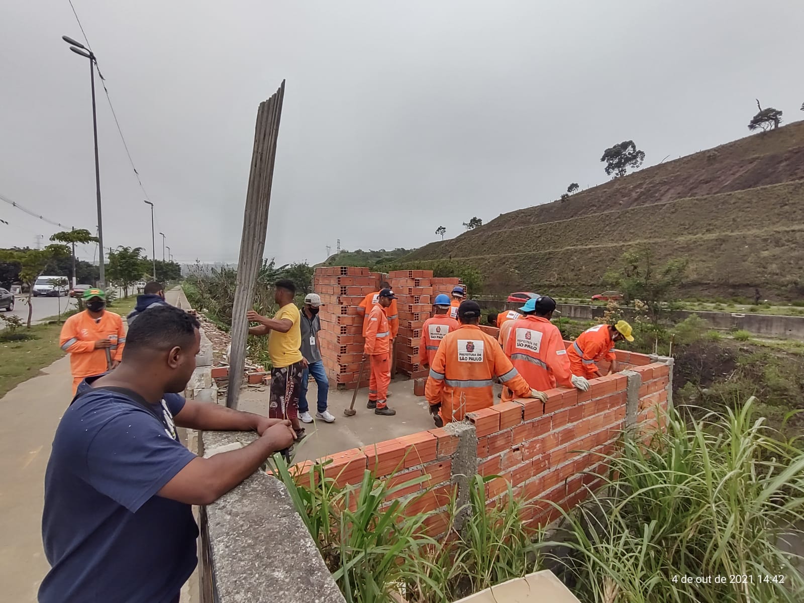 Equipe de funcionários da Prefeitura, com uniforme laranja, trabalham na demolição de paredes de bloco baiano. que estavam sendo erguidas ao lado de um muro de proteção da Avenida Jacu Pêssego. 