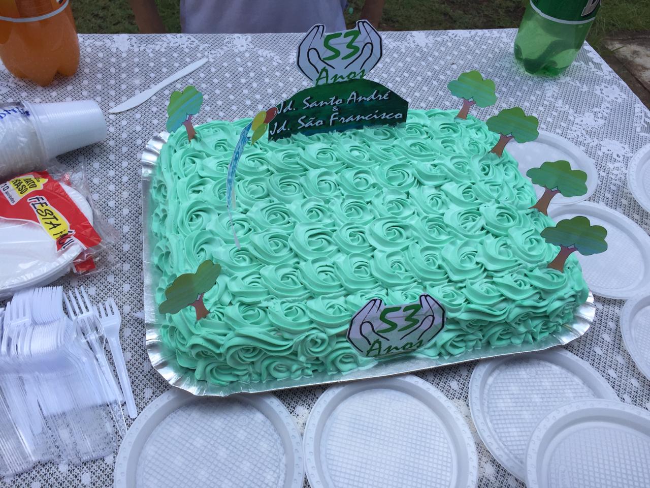 Bolo de aniversário, com glacê na cor verde, placa 53 anos Jardim Santo André e Jardim São Francisco. Miniaturas de árvores foram colocadas nas bordas superiores do bolo. Copos, pratinhos e talheres de plástico estão ao redor do bolo.
