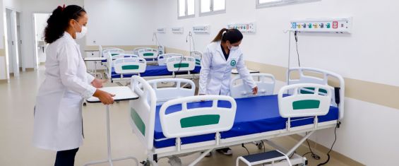 Uma ampla sala de hospital onde duas enfermeiras com uniformes brancos arrumam um dos três leitos para receber pacientes. Os leitos são brancos com colchões azuis.