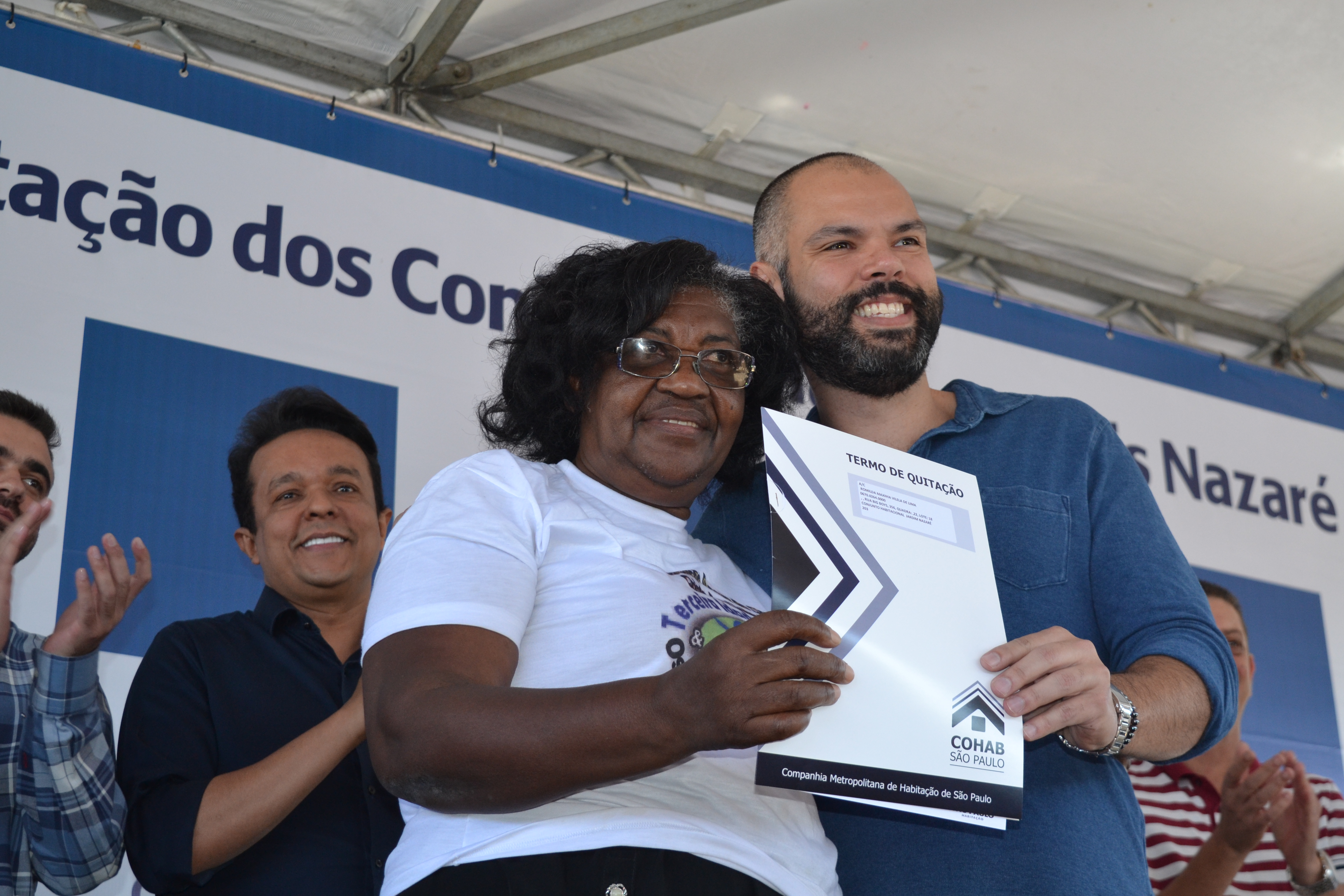 Na foto está o prefeito Bruno Covas com a moradora Romilda segurando o termo de quitação da Cohab. Ao fundo tem algumas autoridades da prefeitura aplaudindo.