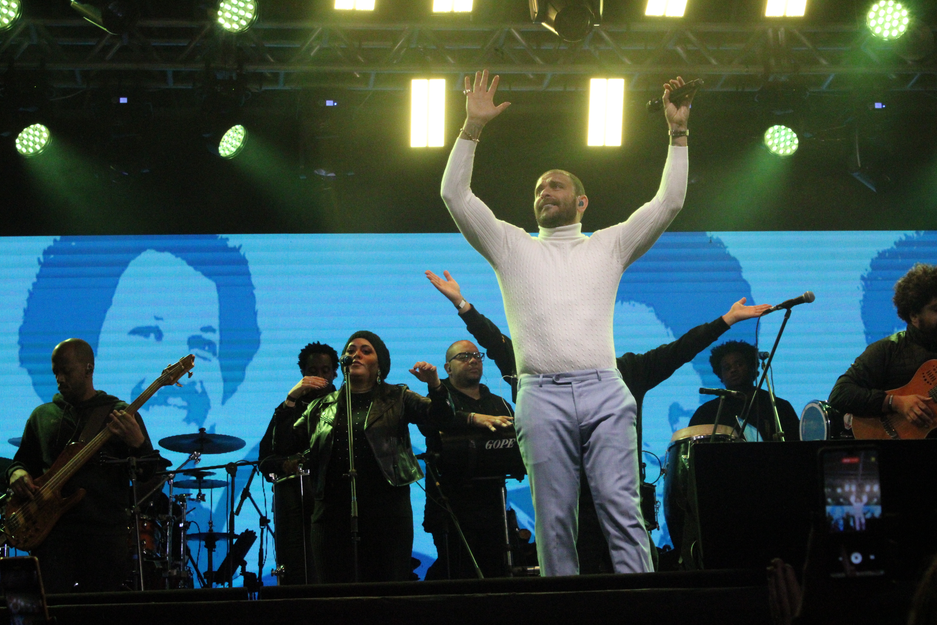 Foto do cantor no centro do palco erguendo os braços, segurando o microfone com a banda atrás.
