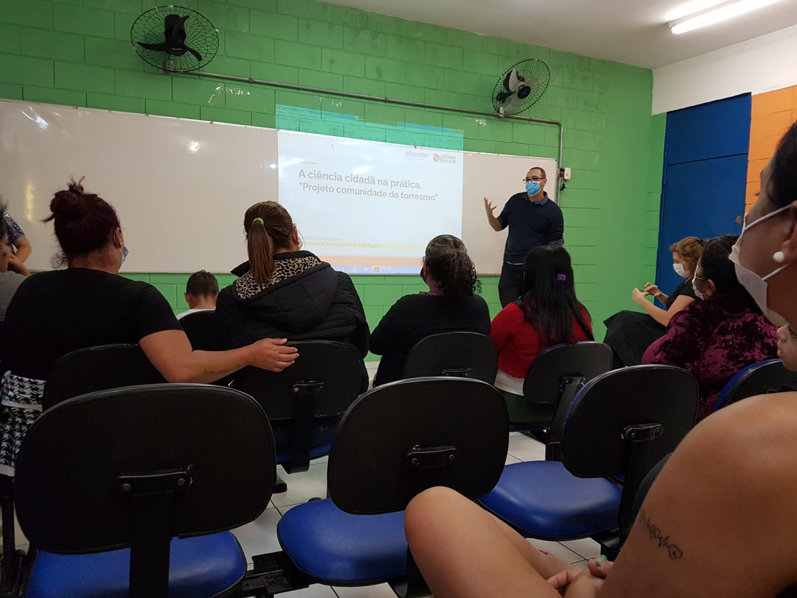 Imagem apresenta pessoas de costas sentadas na sala de aula olhando para um professor que está apresentando na lousa informações do Lab Itaim Paulista.