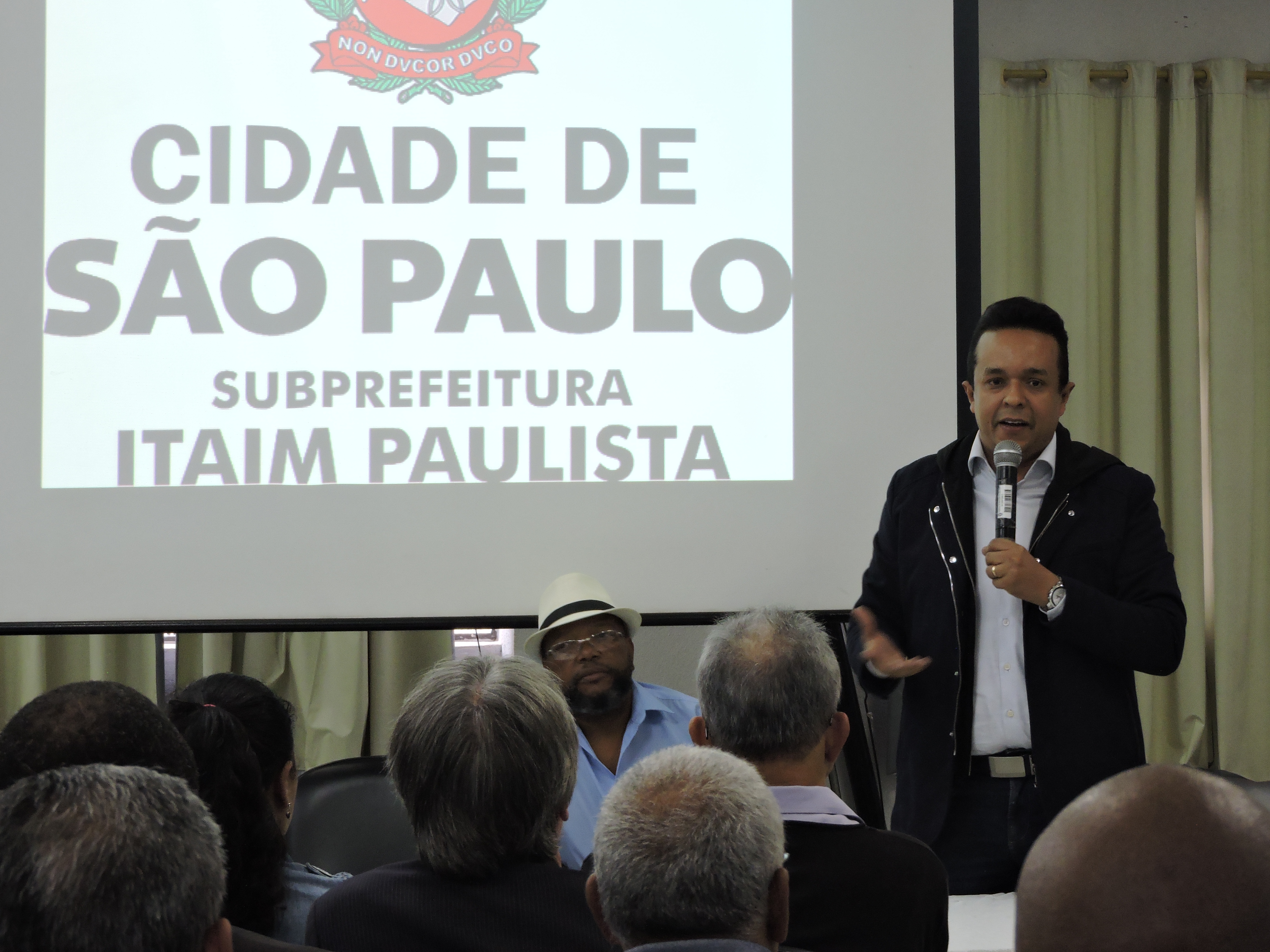 No primeiro plano da foto aparecem cabeças de pessoas sentadas observando o subprefeito Gilmar Souza Santos falando. Ao fundo dele mostra um painel com o logotipo da subprefeitura Itaim Paulista.