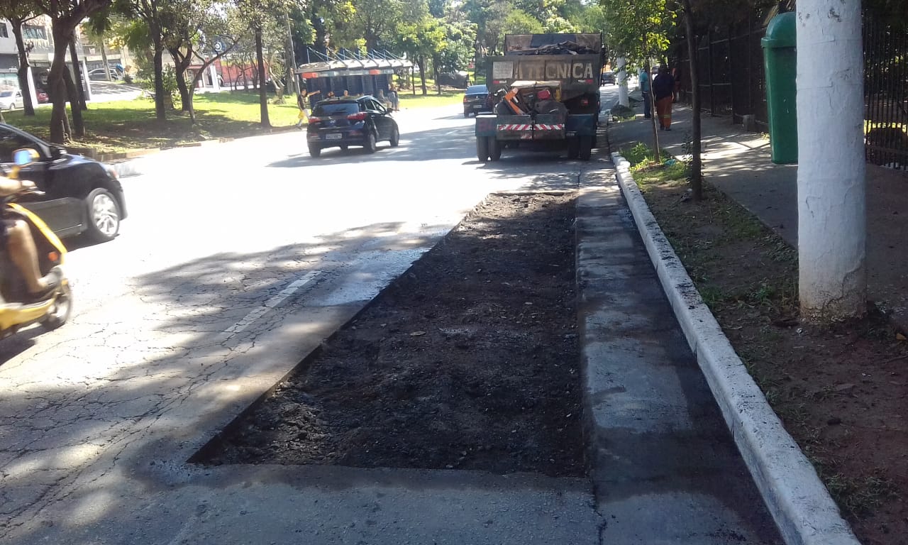 Imagem mostra abertura do asfalto na via na faixa da direita, pronta para receber a massa asfáltica do Tapa-Buracp e o caminhão do serviço ao fundo