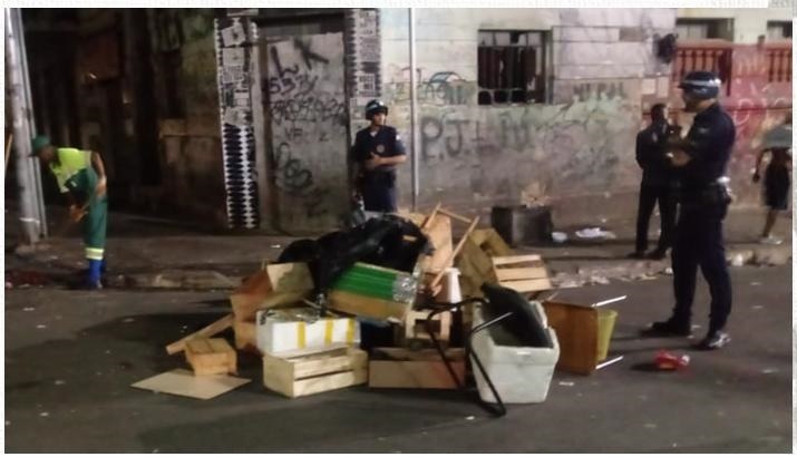 Um funcionário da Subprefeitura Ipiranga varrendo uma ruam no maio da rua tem um monte com caixotes de madeira e três policiais estão acompanhando o serviço.
