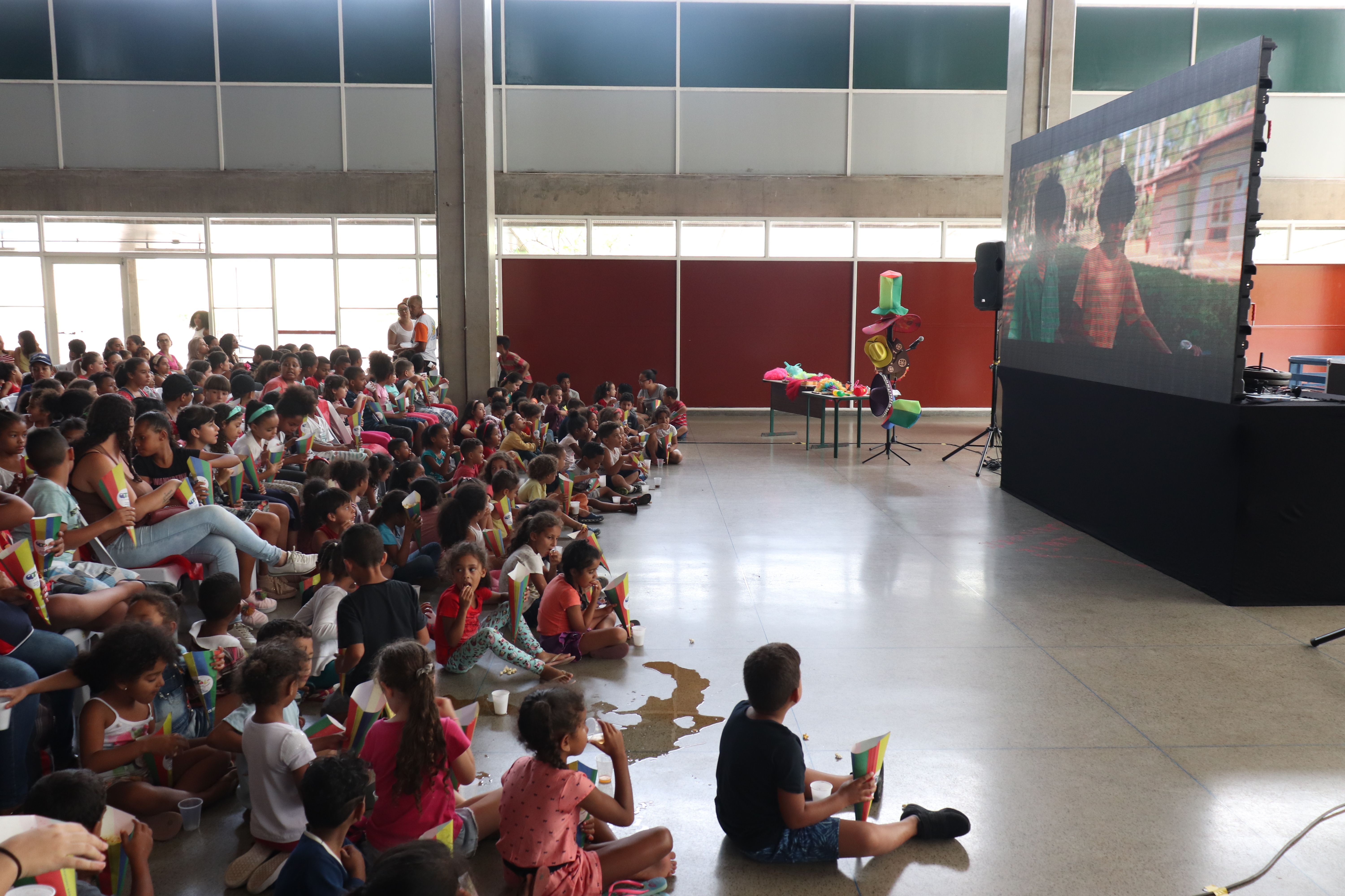 Do lado esquerdo uma plateia repleta de crianças sentadas comendo pipoca e do lado direito um telão exibindo filme” Turma da Mônica: Laços”.