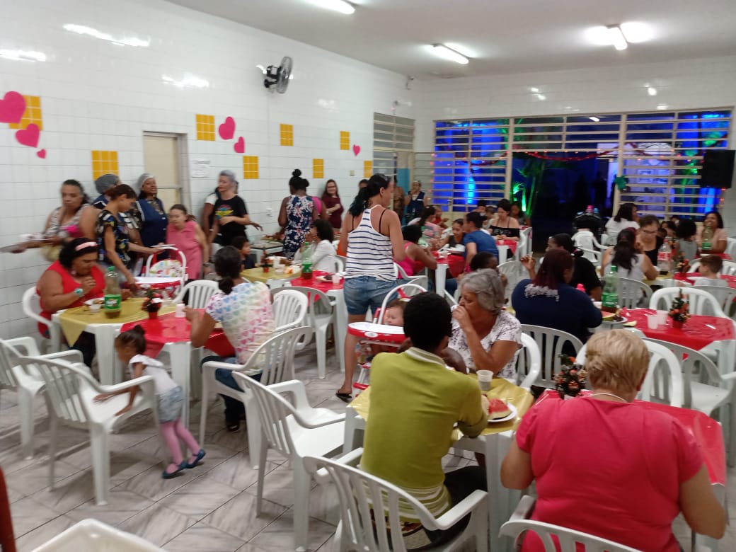 Um salão com várias mesas espalhadas com pessoas participando da ceia de ano natal.