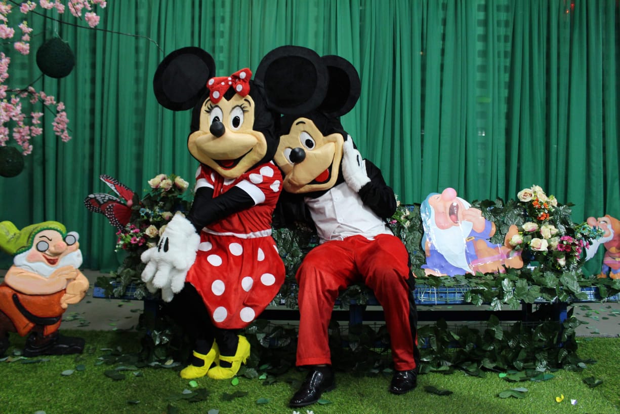 Duas pessoas com fantasias do Mickey e da Minnie sentadas em um banco.