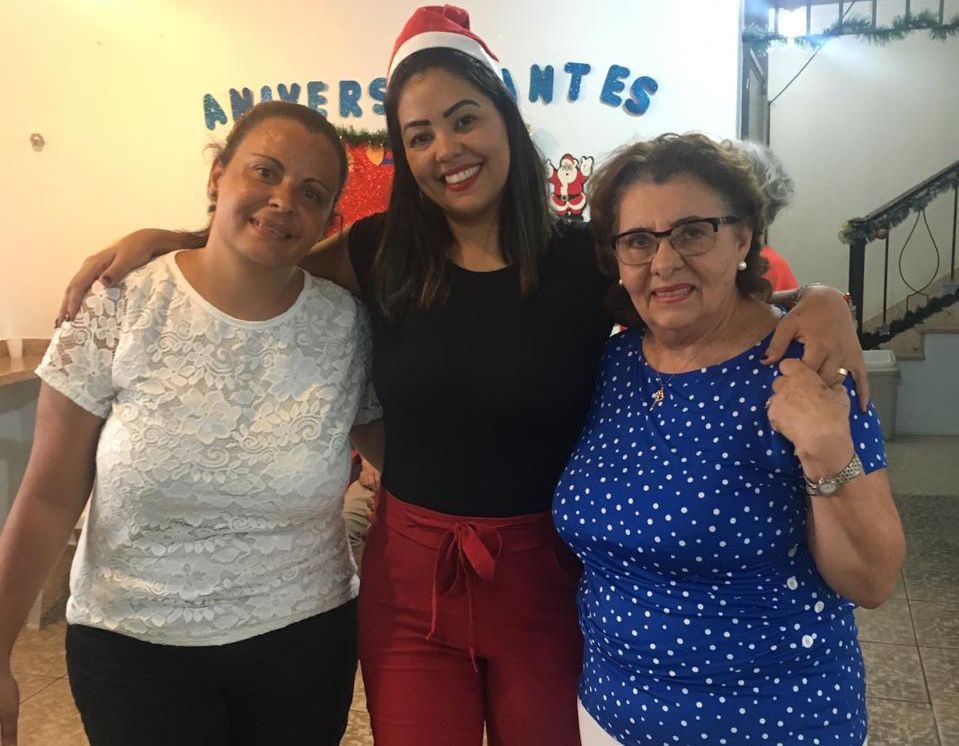 Três mulheres abraçadas posando para a foto. A do centro veste um chapéu de papai Noel