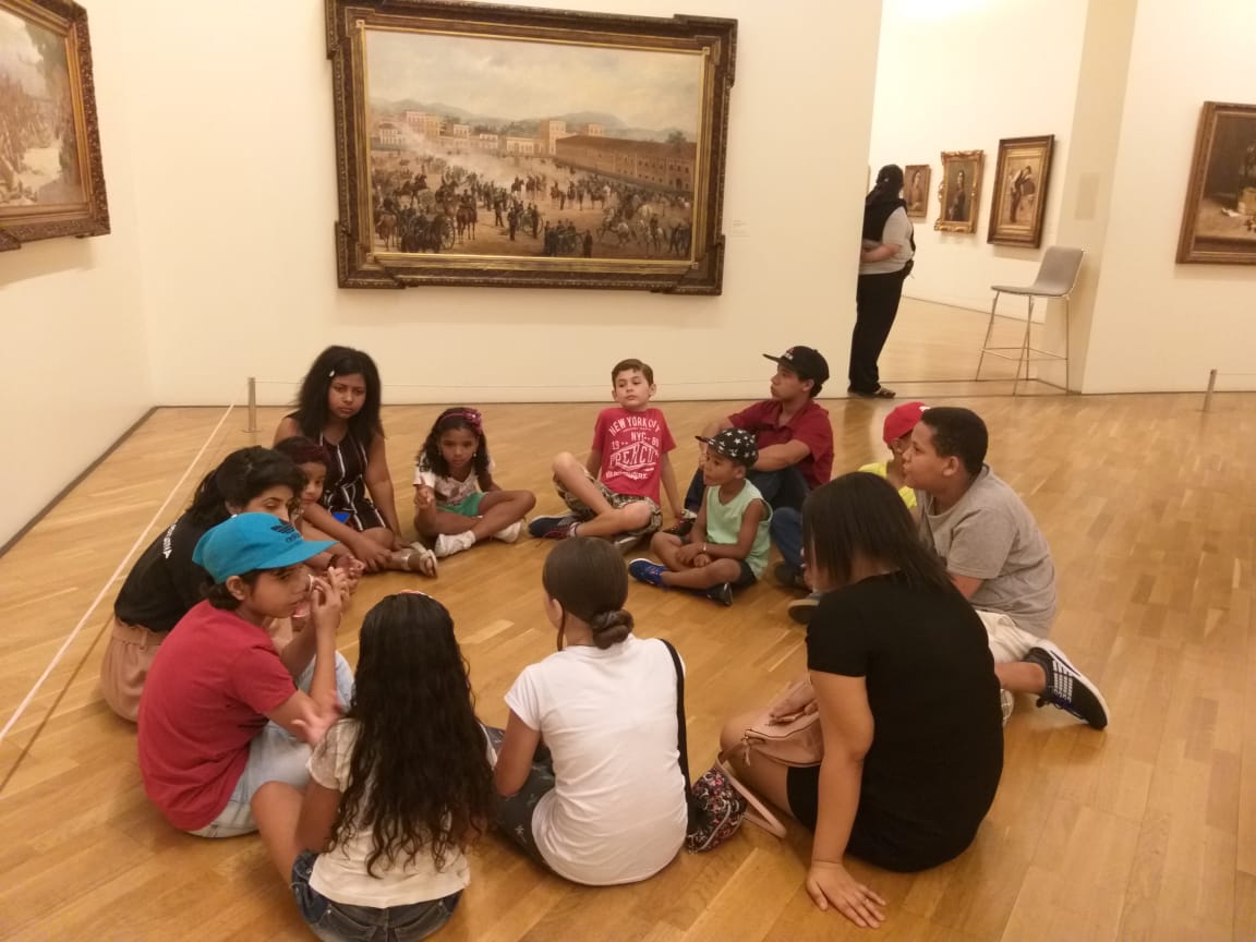 Crianças sentadas em roda no chão de uma sala de exposição.