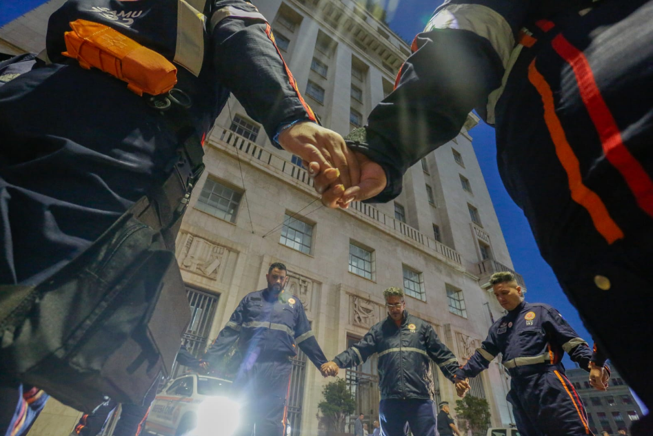A foto mostra profissionais do Samu de mãos dadas em frente ao prédio da Prefeitura