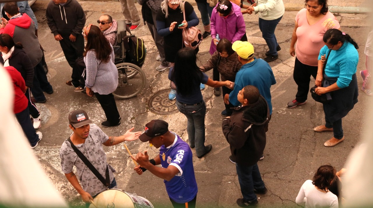 Dezessete pessoas estão dançando e sambando ao som de alguns instrumentos de bateria de escola de samba