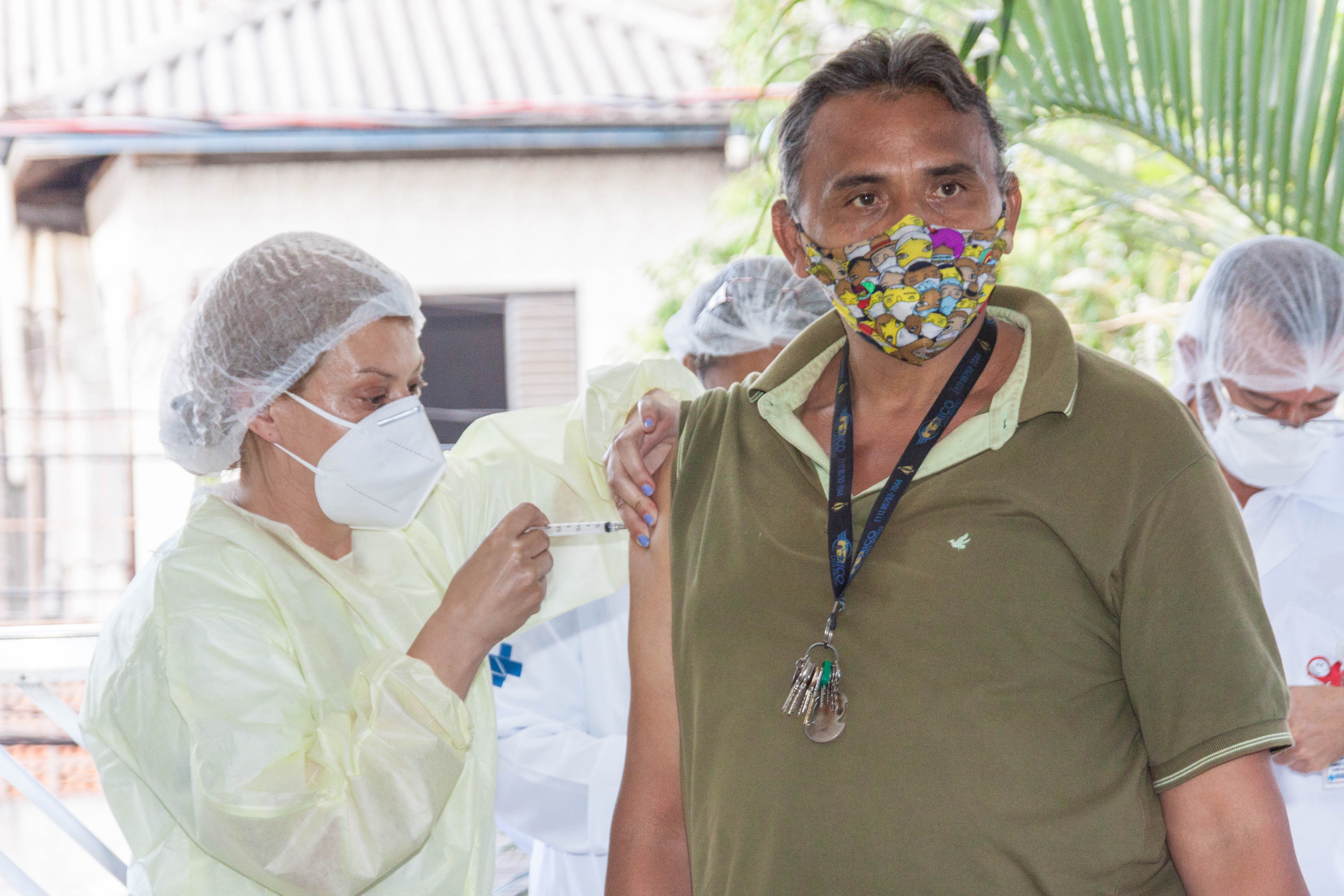 Homem de camiseta verde e uma máscara colorida recebe aplicação de vacina por uma mulher que veste colete, máscara e touca branca.