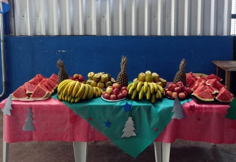 Abacaxis, bananas, maças, melancias e kiwis são servidos em três mesas brancas cobertas com toalhas vermelhas e verdes e pequenas ilustrações de árvores de Natal.