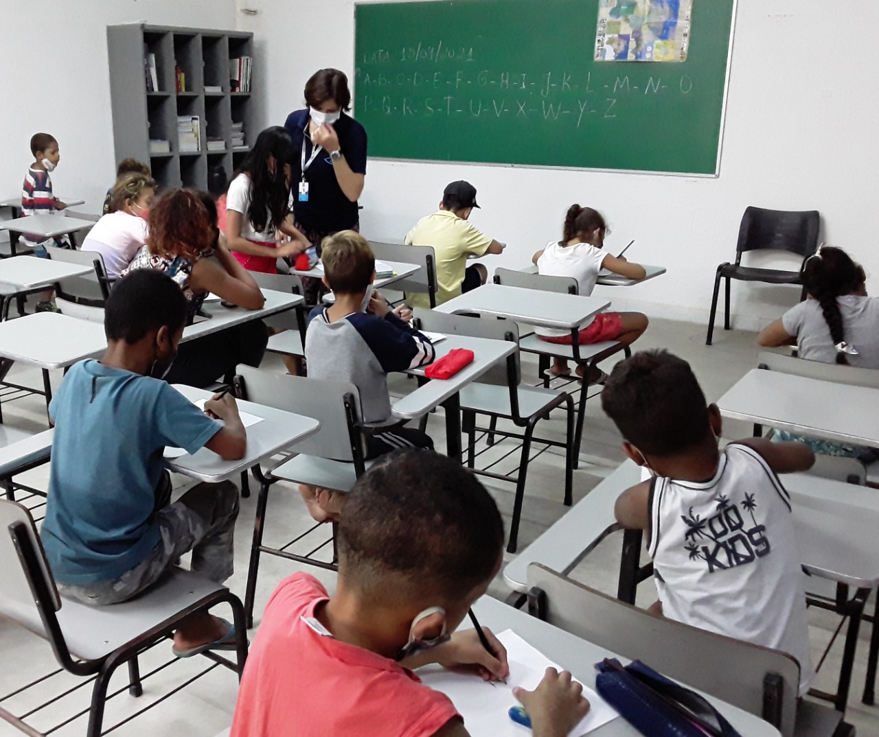 Em uma sala de aula, crianças com máscaras de proteção facial estão sentadas escrevendo, há uma professora na sala explicando o conteúdo. Na lousa está escrito o alfabeto. 