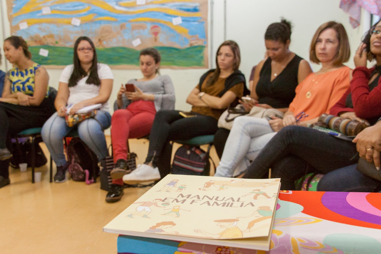 A imagem destaca um manual, que está sob um bancada com o título escrito na capa"Manual Família". Atrás, aparecem sete mulheres sentadas em carteiras escolares, dentro de uma sala de aula.