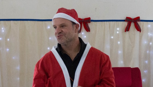Barrichello, vestido com gorro e roupa de Papai Noel por cima de uma camiseta preta, tira a barba que compõe o traje para se apresentar às crianças. O piloto está sorrindo, com o rosto levemente de perfil.