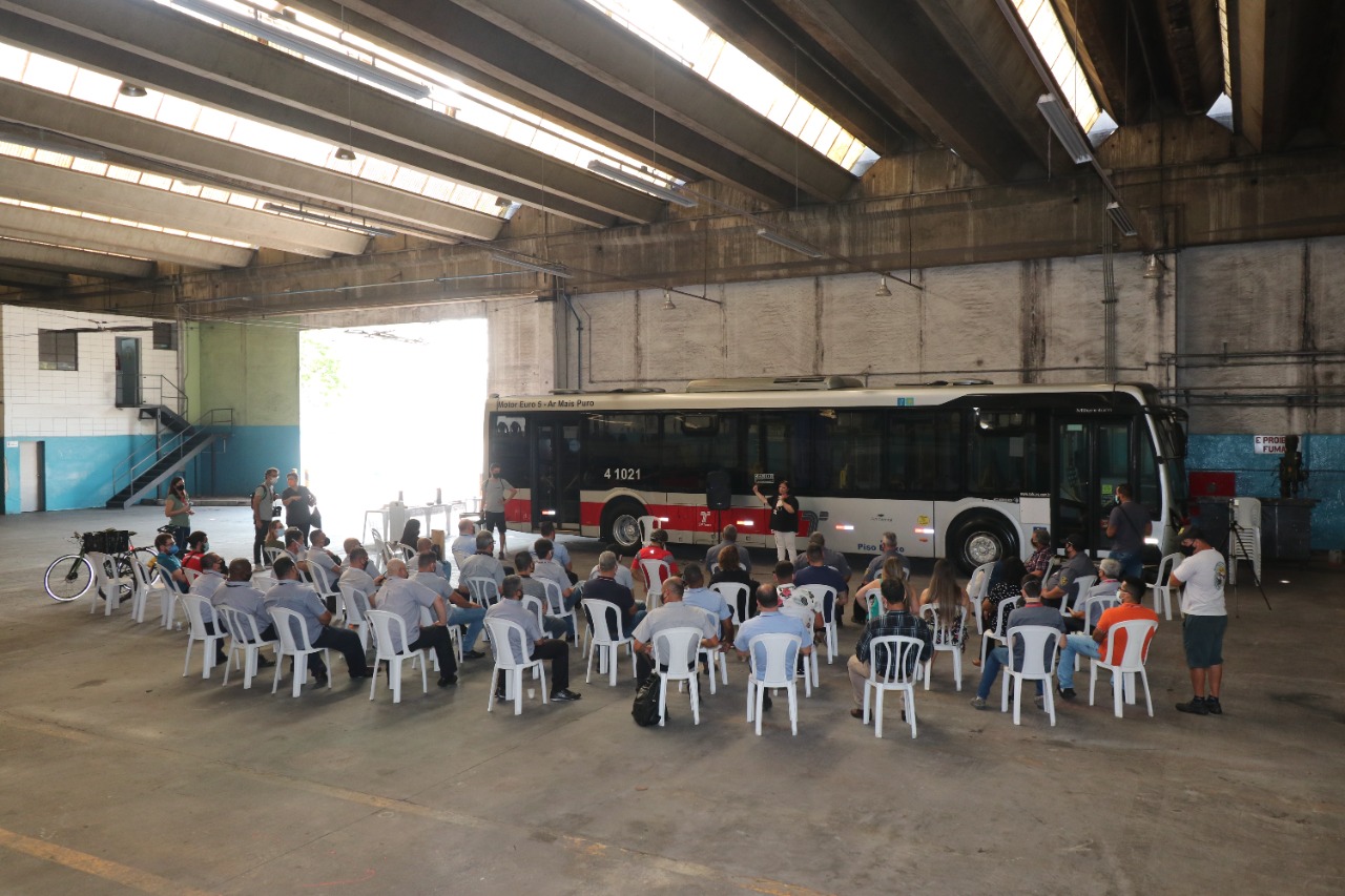 Imagem de um galpão com um ônibus e dezenas de motoristas sentados e ouvindo a palestra