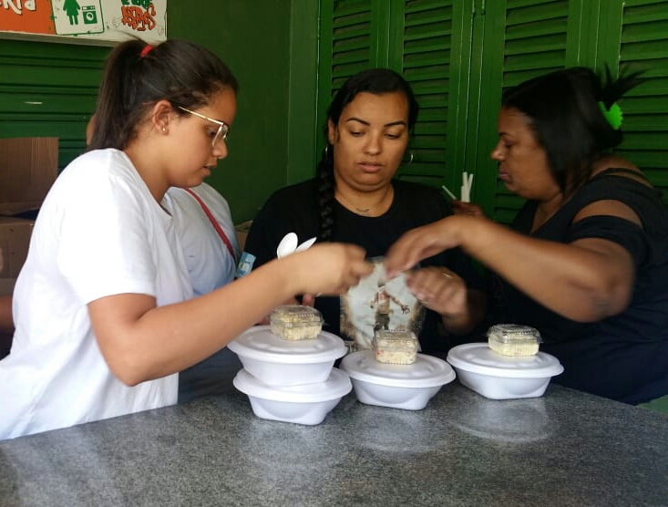 Três funcionárias do serviço fecham marmitas de isopor com a comida dos usuários ao redor de uma mesa