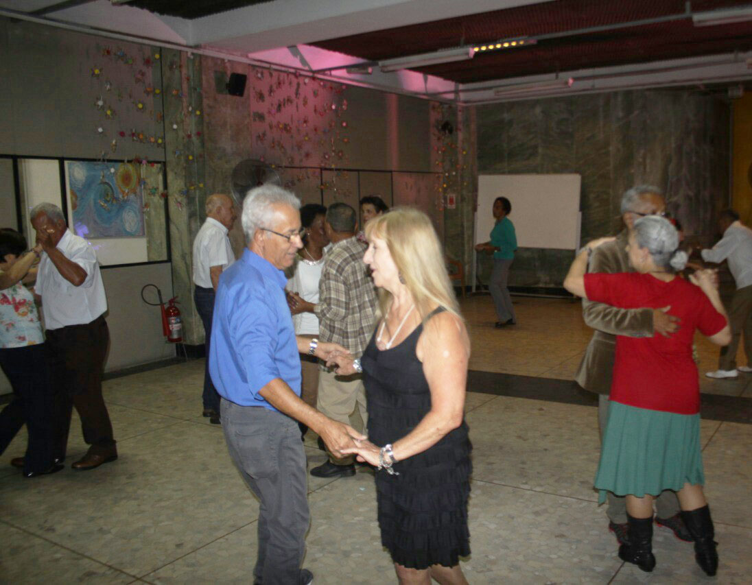 Casais dançam espalhados pelo salão enquanto uma mulher dança sozinha no centro da foto