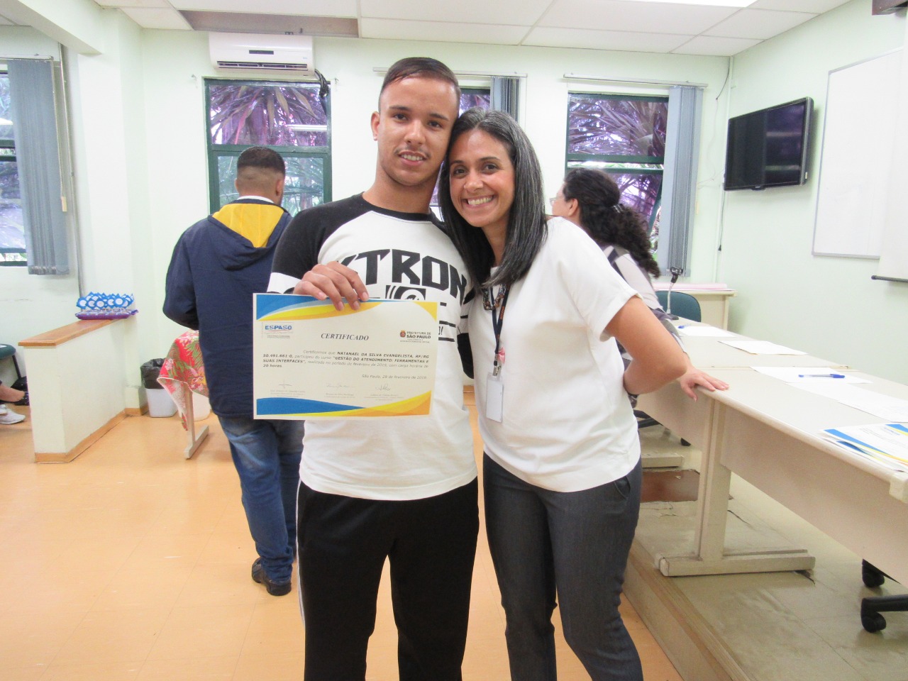 Formando com diploma na mão ao lado da supervisora do ESPASO, Lidiane Borges, ambos em pé na sala de aula. 
