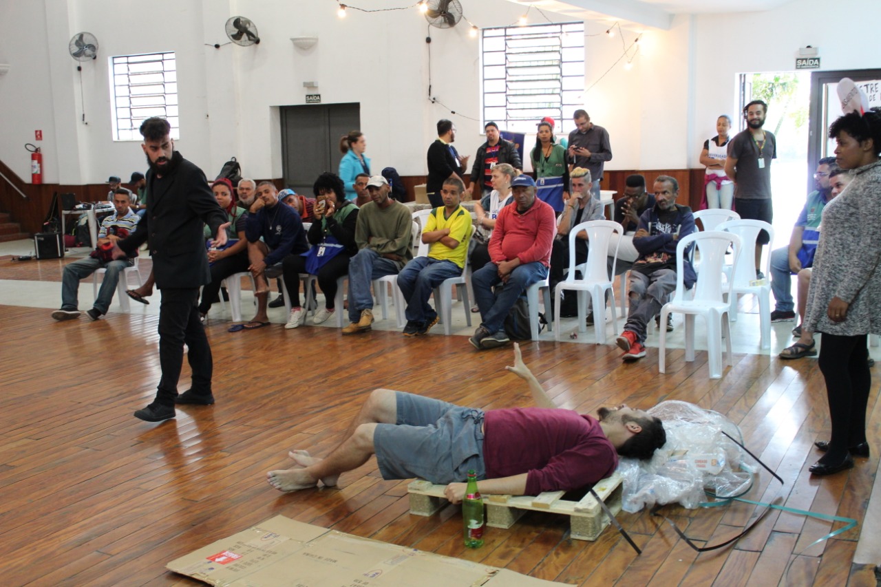 Um ator deitado no chão contracena com ator em pé diante de uma plateia sentada ao fundo