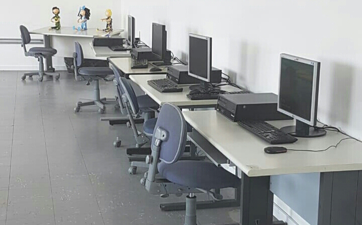 Sala de informática vazia com quatro computadores e suas cadeiras, além de uma cadeira ao fundo com mesa onde se vê três bonecos coloridos em cima