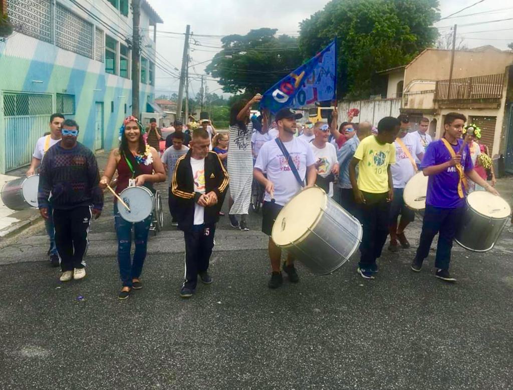 Conviventes caminham em bloco de Carnaval com cinco integrantes tocando tambor e uma bandeira azul sendo erguida ao fundo com dizeres não visíveis 