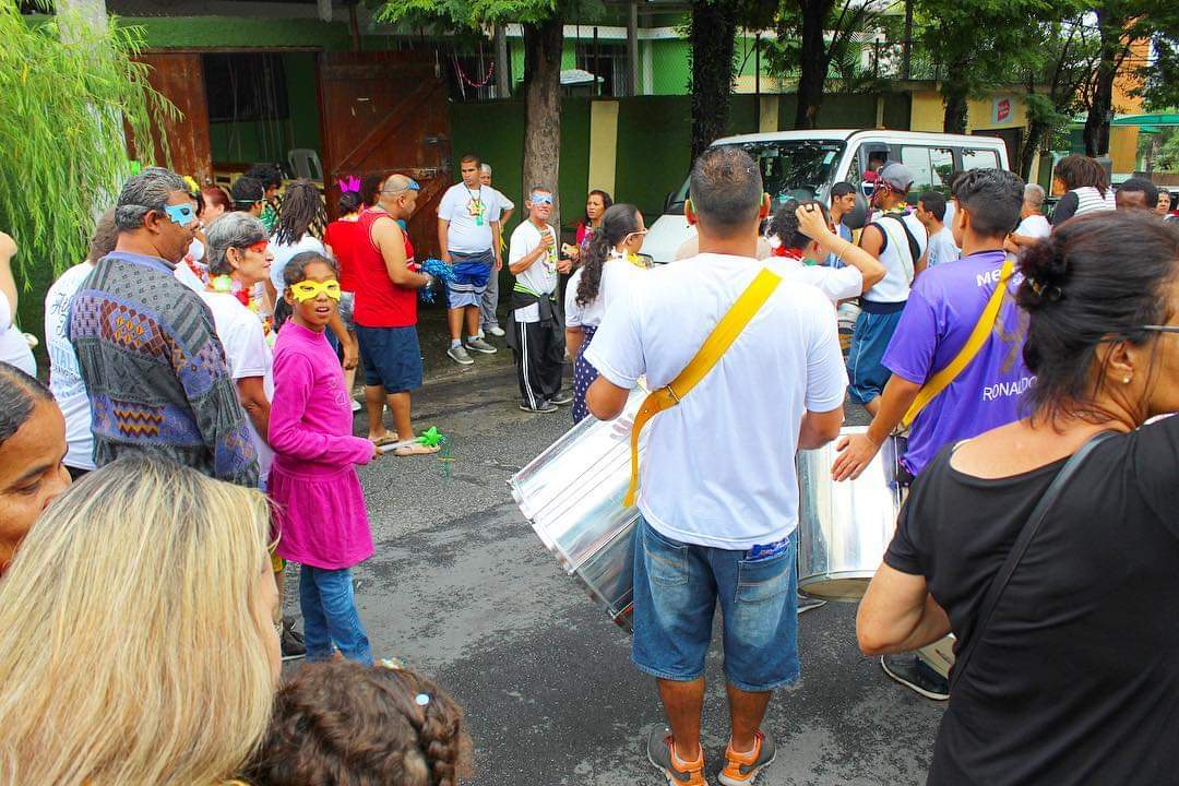 Concentração de conviventes na frente do serviço, incluindo um homem de costas tocando tambor, algumas crianças com máscaras de Carnaval e uma van branca parada ao fundo.