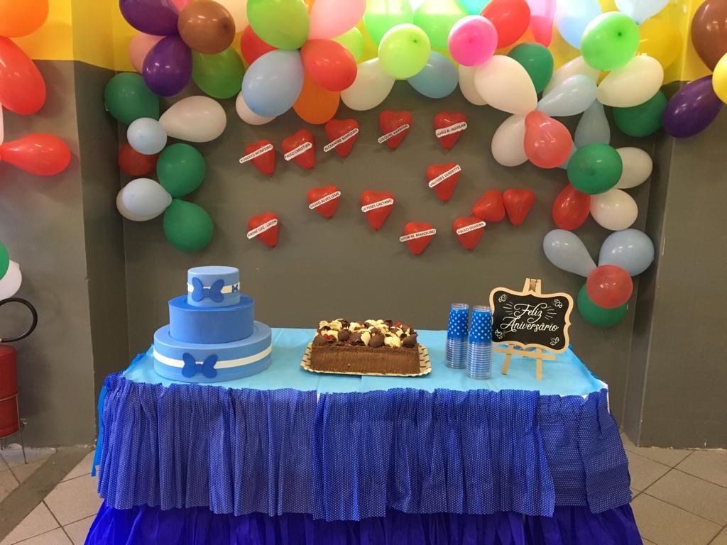 Foto de mesa de aniversário com pano azul, balões coloridos nas paredes ao redor, um bolo à esquerda, azul, de três andares, um bolo confeitado ao centro, copos de plástico empilhados e uma plaquinha de Feliz Aniversário