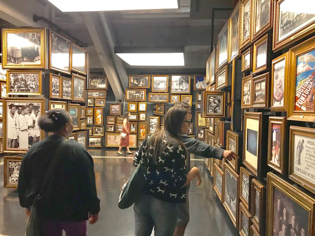 Três jovens olham fotos espalhados em sala de fotografias emolduradas em dourado do chão ao teto