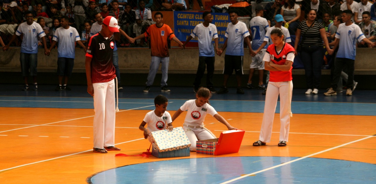 Duas crianças abrem caixas coloridas, agachadas no centro da arena de futsal, enquanto uma menina e um menino olham em pé, um de cada lado, para as caixas no chão. Ao fundo, conviventes um ao lado do outro, de mãos dadas, vestindo uniforme