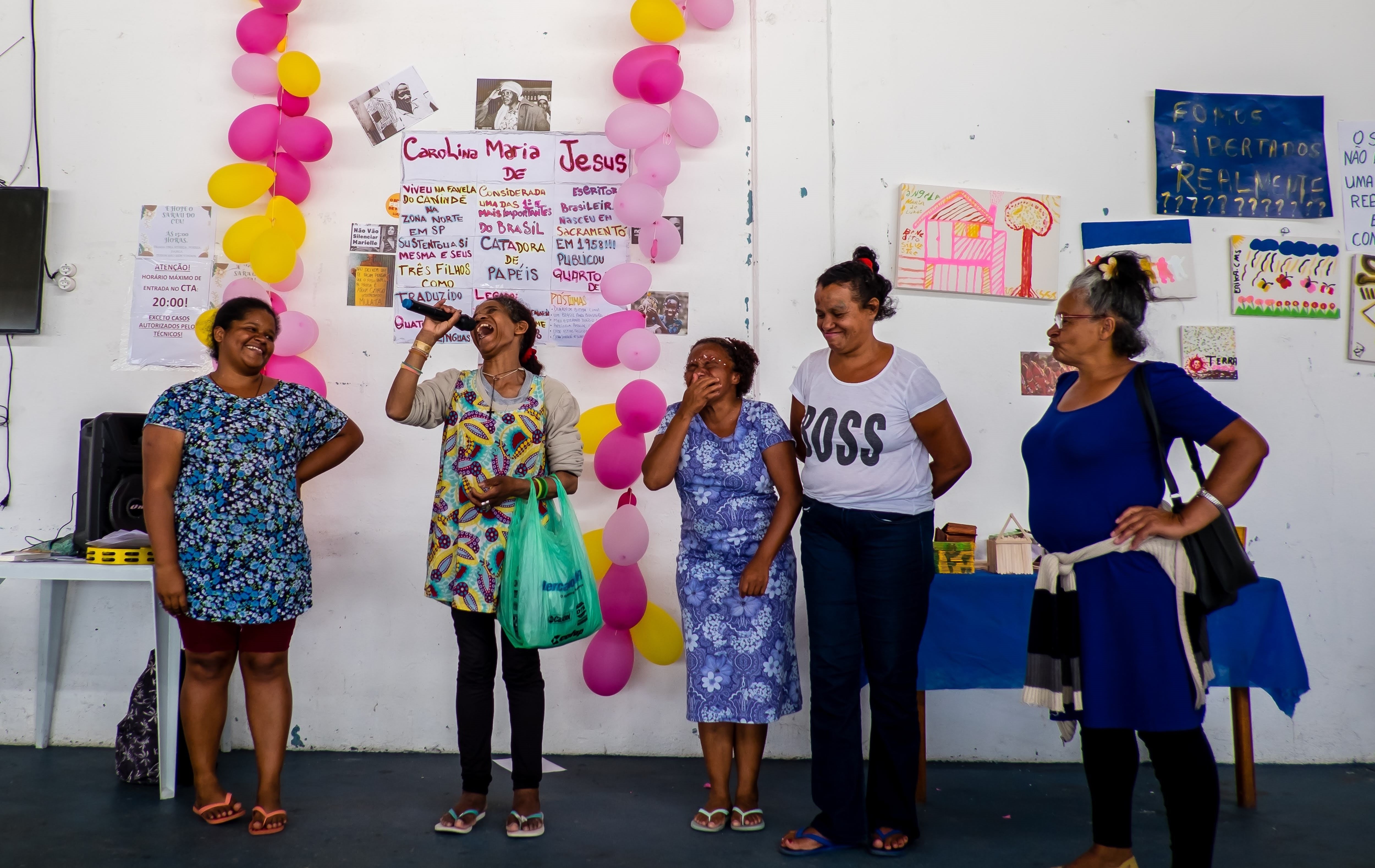 Cinco conviventes no palco rindo enquanto uma delas canta ao microfone com balões amarelos e rosas na parede dos fundos