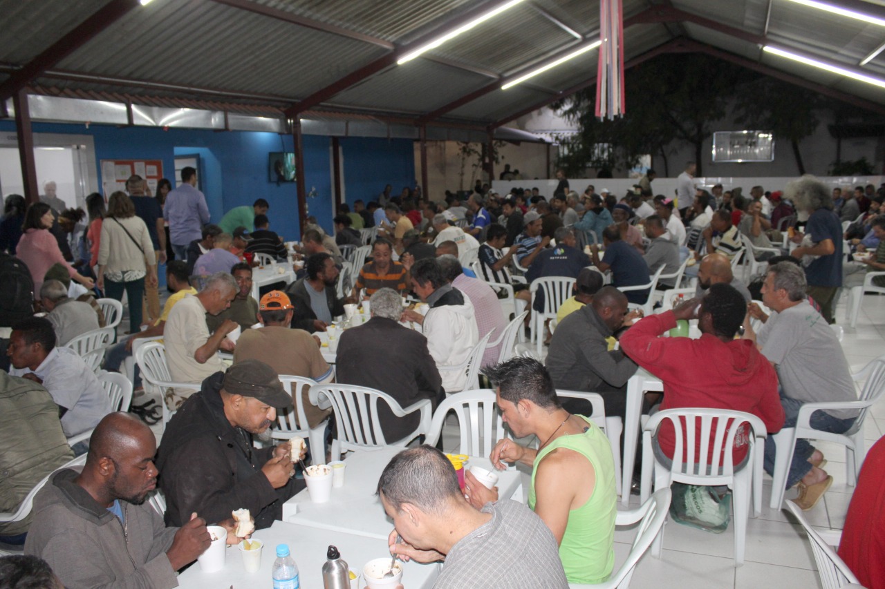 Mais de cem conviventes jantam em mesas brancas de plástico no refeitório.