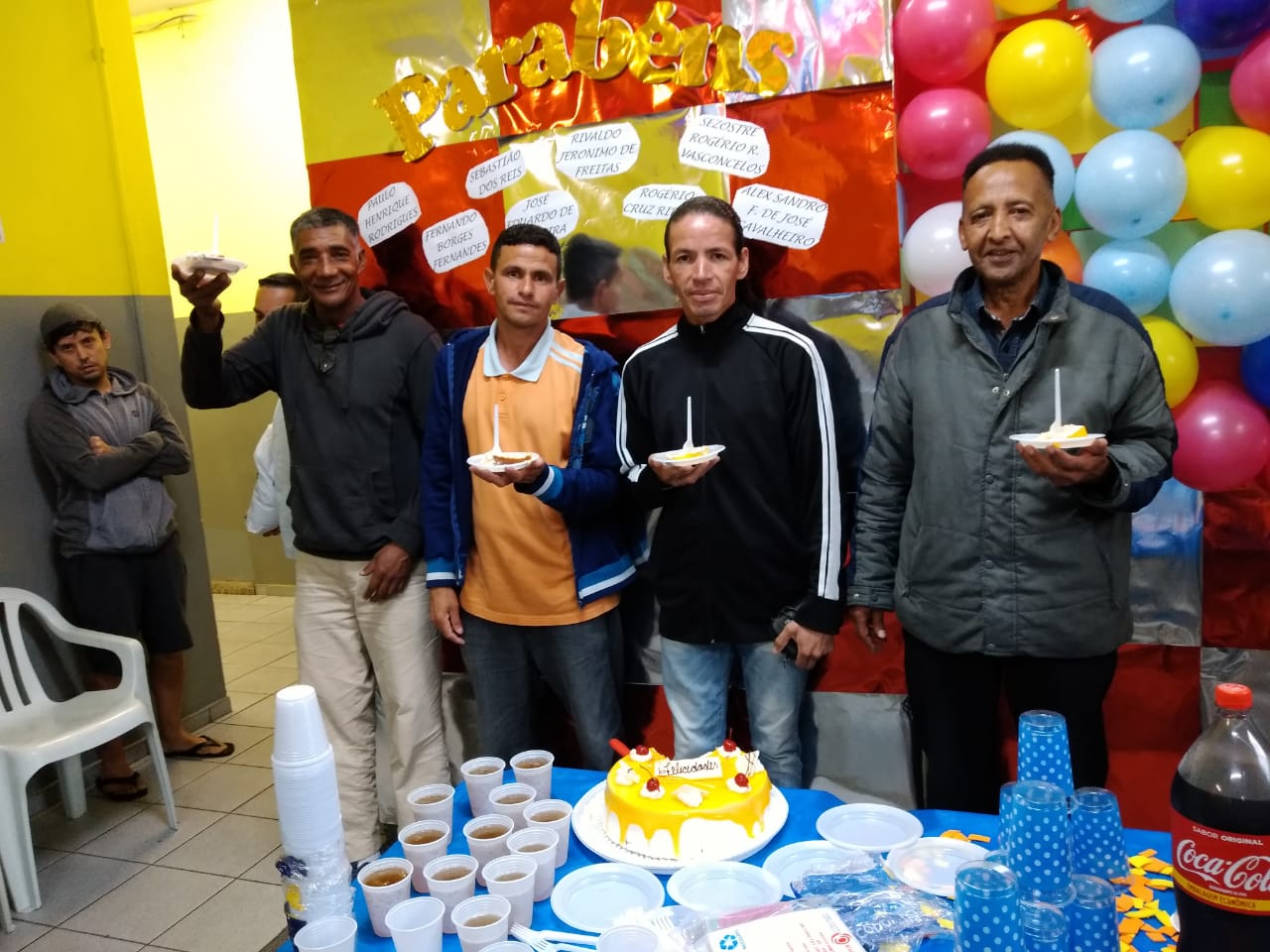 Quatro homens sorrindo para a foto, seguram pratos com bolo e estão em frente a mesa de aniversário onde está o bolo, copos, pratos e refrigerante. Ao fundo, painel de bexigas coloridas. 
