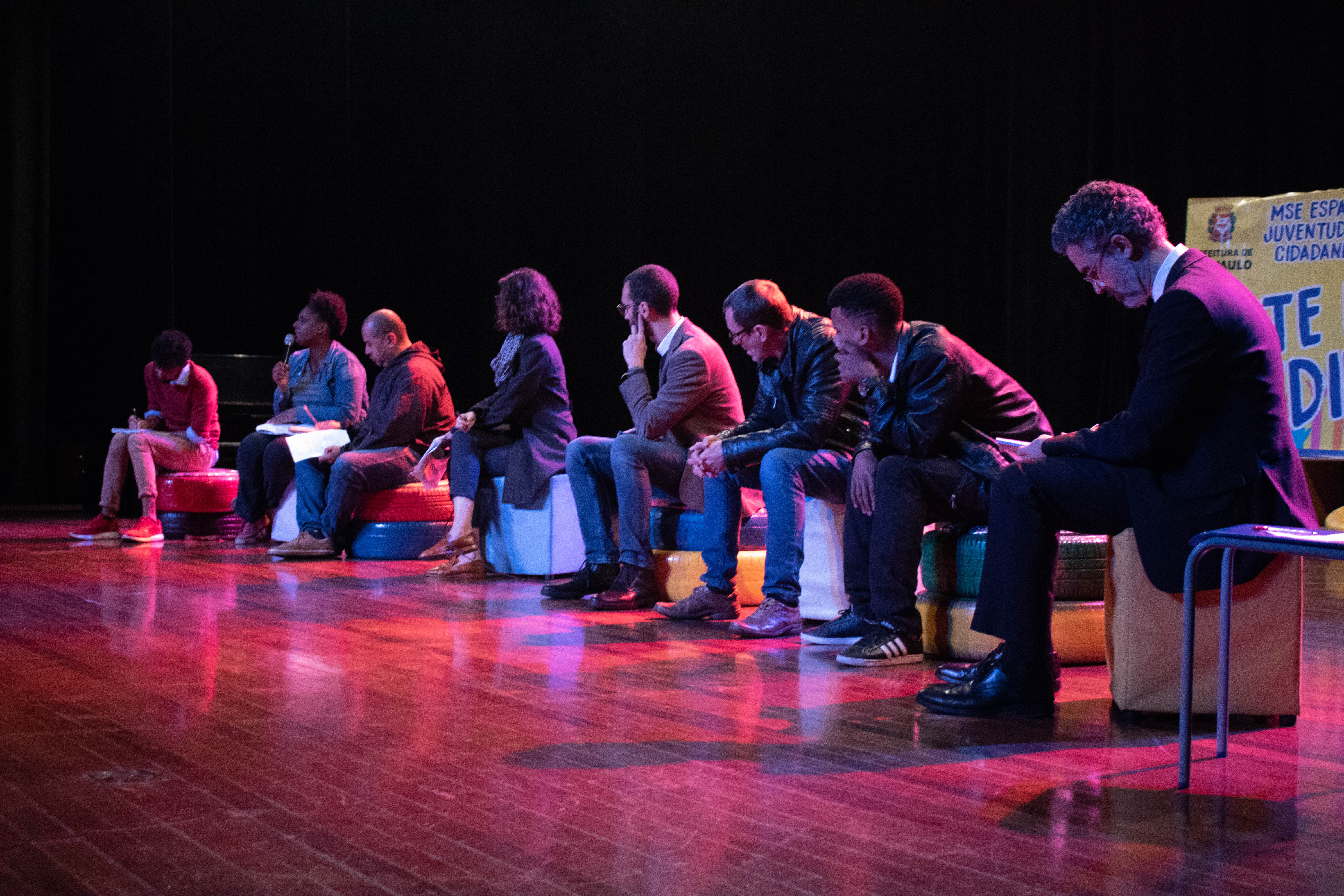 Oito palestrantes sentados no palco com um painel ao fundo escrito “Arte na medida”.