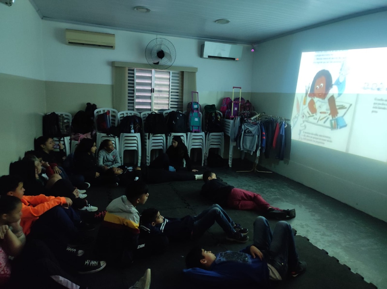 Crianças sentadas no chão de uma sala escura observam a tela do projetor, que ilustra páginas do livro. Todas estão como olhar fixo para a tela. 