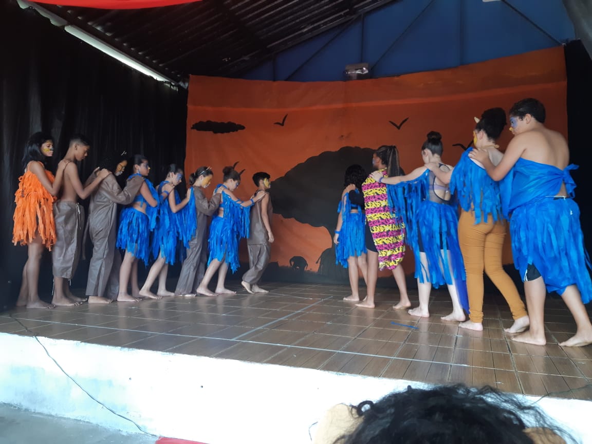 Treze adolescentes estão em cima de um palco interpretando uma peça do Rei Leão. Eles usam roupas na cores azul e cinza.