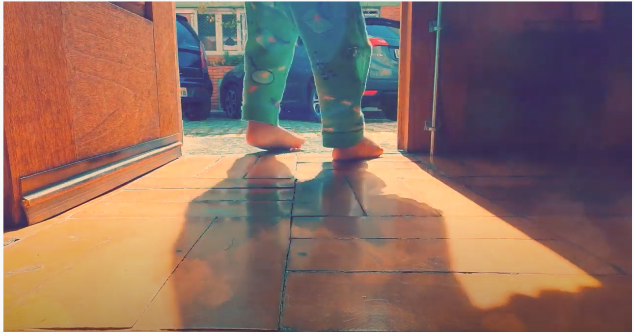 Foto mostrando os pés do bebê descalço em frente a porta aberta com piso de madeira e fundo de tons marrons.