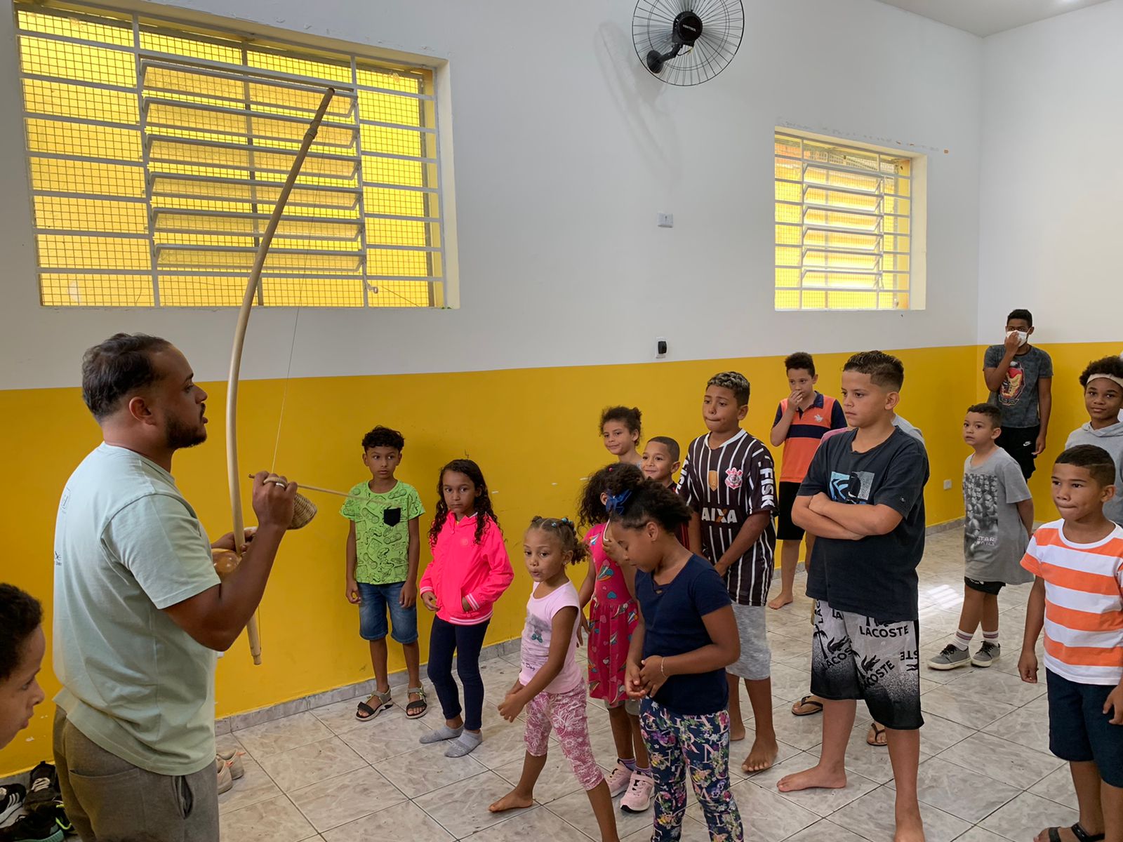 Na foto, algumas crianças estão organizadas uma ao lado da outra, em uma atividade sobre capoeira. Na frente delas, um homem segura um berimbau. Ao fundo tem duas janelas e um ventilador, em uma parede branda a amarela.