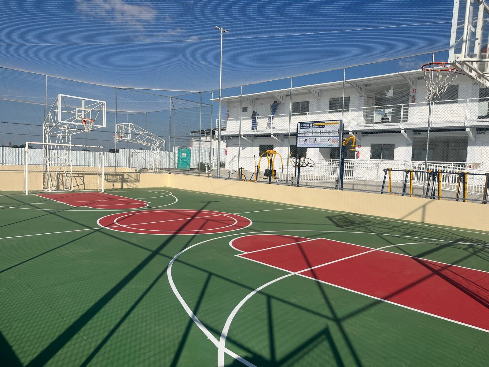 Na imagem aparece a quadra poliesportiva da Vila Reencontro Jabaquara. O chão está pintado de verde, vermelho e branco. Ao fundo módulos administrativos do serviço.