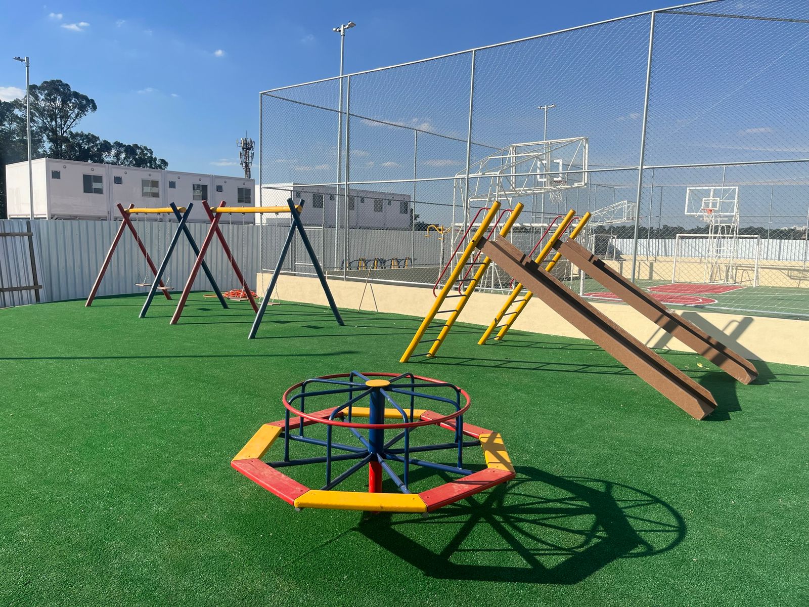 Na imagem aparece o playground da Vila Reencontro. Na imagem aparecem um gira-gira colorido, dois escorregadores é um balanço