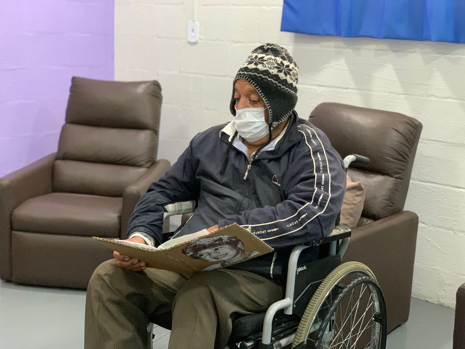 Homem idoso sentado em uma cadeira de rodas com um livro aberto sobre o colo.