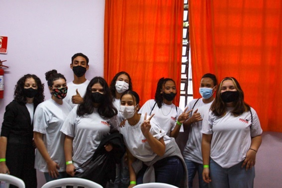 Nove Jovens do CJ Mutirão vestidos com o uniforme, todos utilizam mascaras, e estão na frente de uma cortina vermelha.