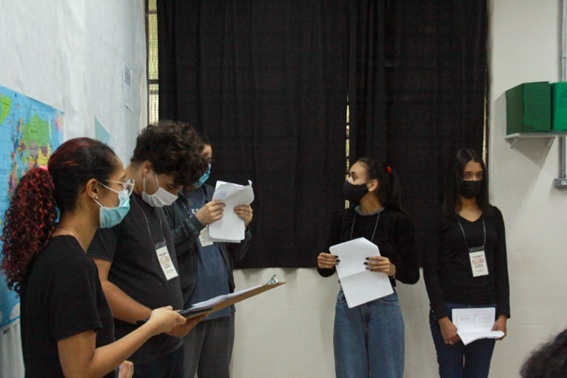 cinco pessoas presentes na imagem, todas estão vestidas de preto e de máscaras, e seguram em suas mãos um papel para a realização da apresentação. 