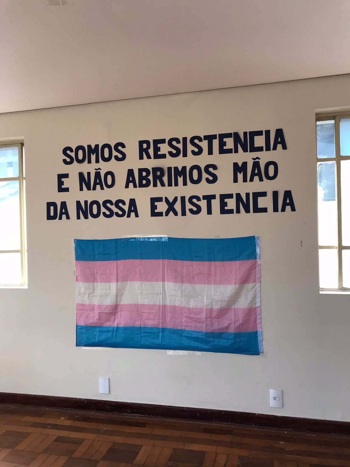 Foto da parede decorada com uma frase escrita em preto “Somos resistência e não abrimos mão da nossa existência” e logo a baixo a bandeira de representatividade trans 