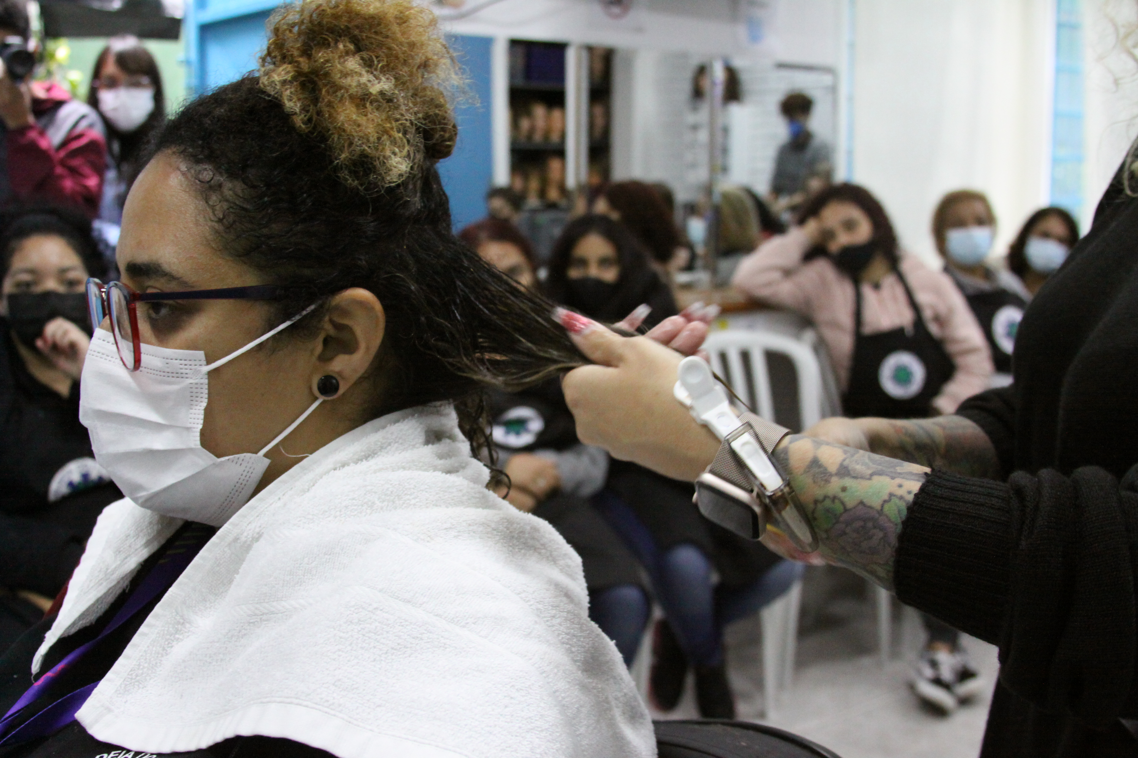 Voluntaria mostrando o procedimento de finalização em aluna que está de mascará e óculos. Ai fundo as alunas de mascará observam a técnica